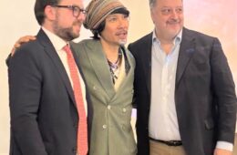 Three men in business attire posing together at an event, middle person wearing a striped headscarf and green double-breasted blazer.