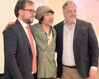 Three men in business attire posing together at an event, middle person wearing a striped headscarf and green double-breasted blazer.