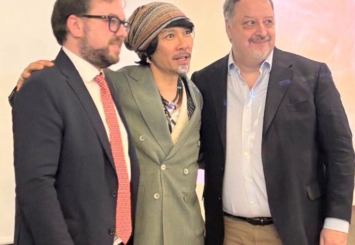 Three men in business attire posing together at an event, middle person wearing a striped headscarf and green double-breasted blazer.