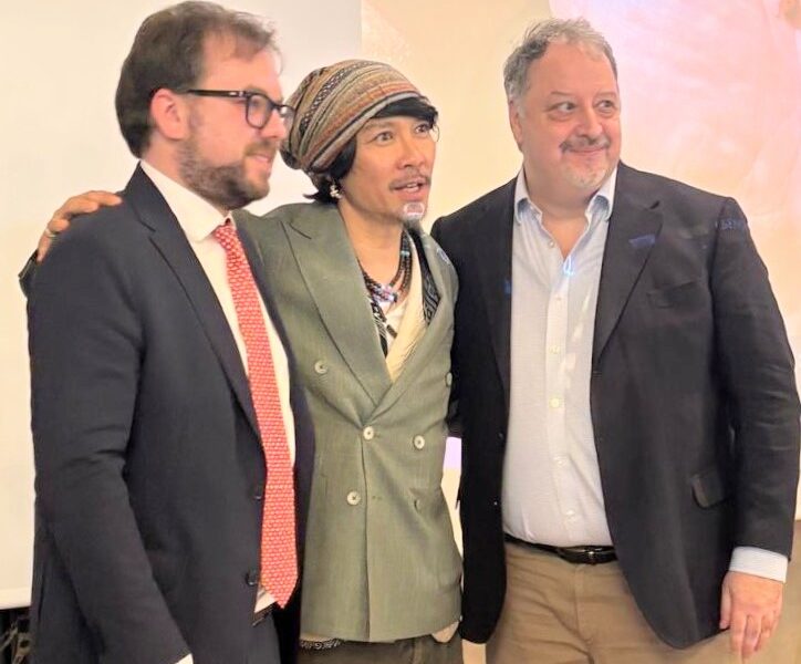Three men in business attire posing together at an event, middle person wearing a striped headscarf and green double-breasted blazer.