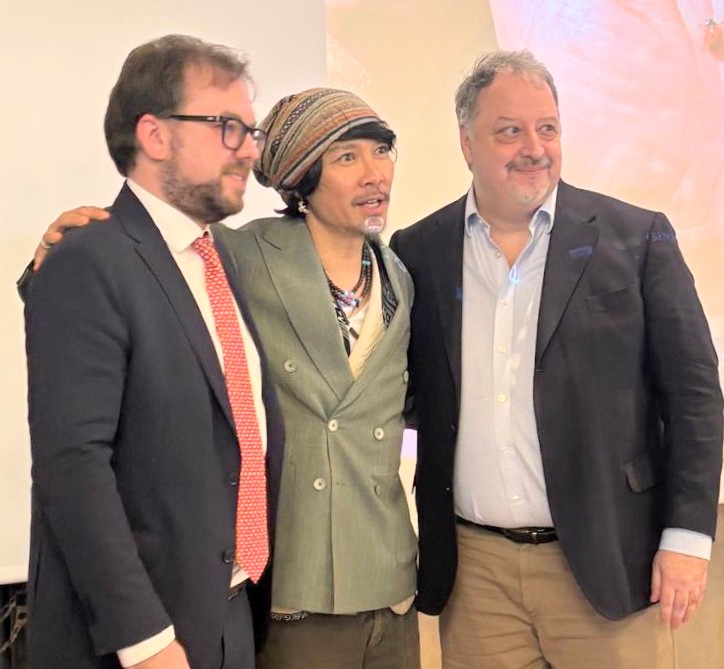 Three men in business attire posing together at an event, middle person wearing a striped headscarf and green double-breasted blazer.