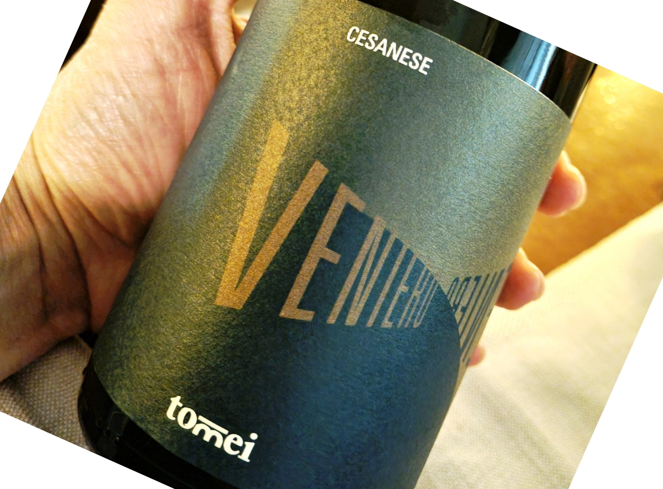 Close-up of a hand holding a dark blue travel mug with gold geometric lines and partially visible white text on the label; fabric background is blurred in the scene.