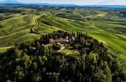 Hilltop rural estate with red-tiled buildings surrounded by cypress trees on a green hillside, with rolling fields and a winding road below the peak