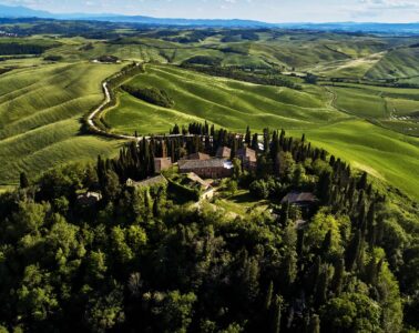Hilltop rural estate with red-tiled buildings surrounded by cypress trees on a green hillside, with rolling fields and a winding road below the peak