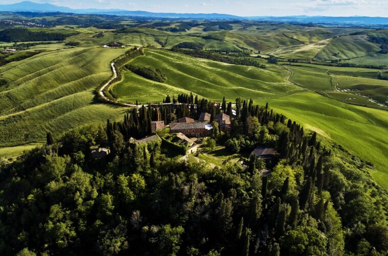 Hilltop rural estate with red-tiled buildings surrounded by cypress trees on a green hillside, with rolling fields and a winding road below the peak