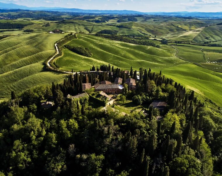 Hilltop rural estate with red-tiled buildings surrounded by cypress trees on a green hillside, with rolling fields and a winding road below the peak