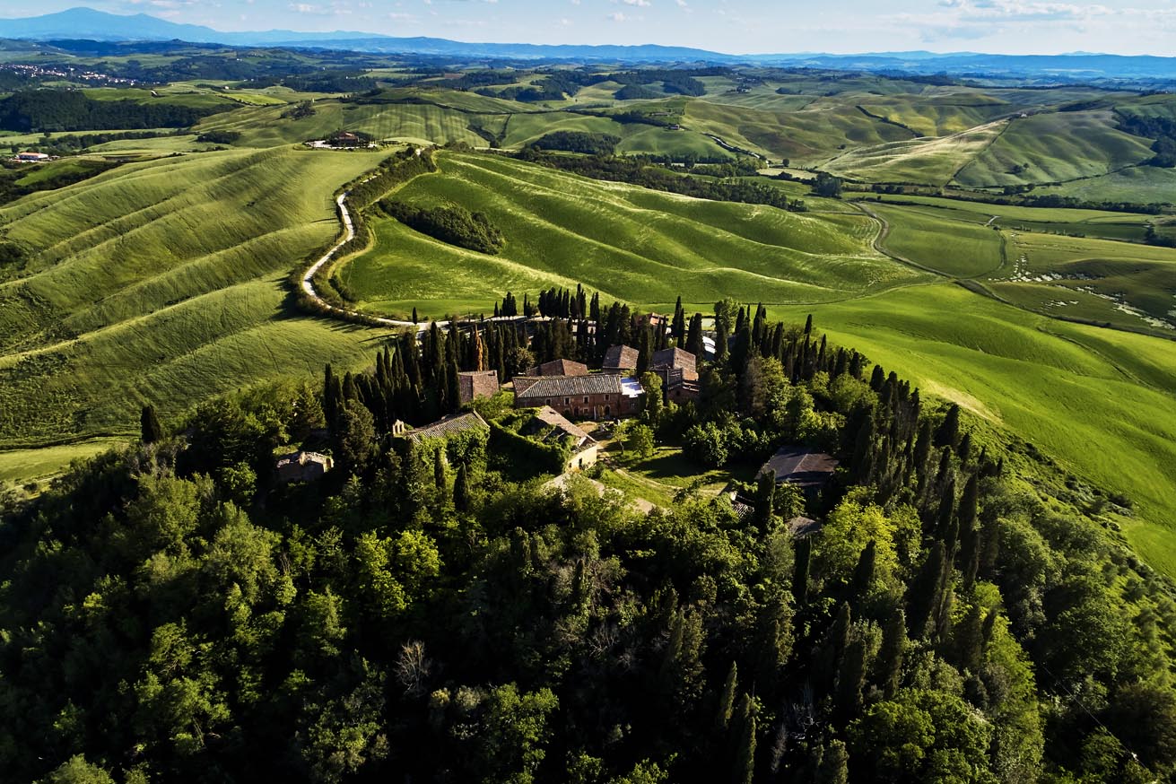 Hilltop rural estate with red-tiled buildings surrounded by cypress trees on a green hillside, with rolling fields and a winding road below the peak