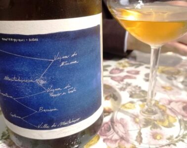Wine bottle with a blue label featuring a handwritten constellation-style map next to a wine glass on a floral tablecloth. The setting appears to be a casual wine tasting or dining scene.