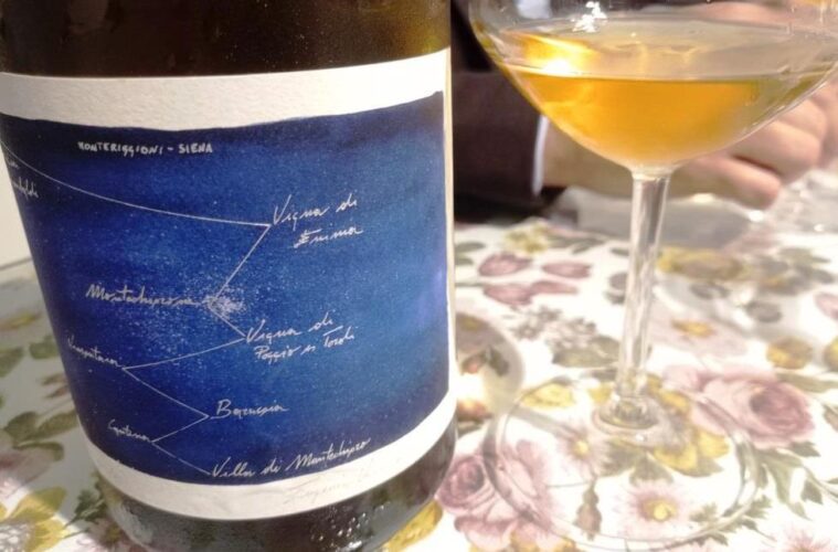 Wine bottle with a blue label featuring a handwritten constellation-style map next to a wine glass on a floral tablecloth. The setting appears to be a casual wine tasting or dining scene.