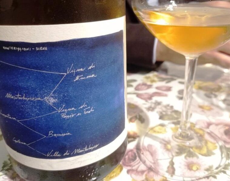 Wine bottle with a blue label featuring a handwritten constellation-style map next to a wine glass on a floral tablecloth. The setting appears to be a casual wine tasting or dining scene.