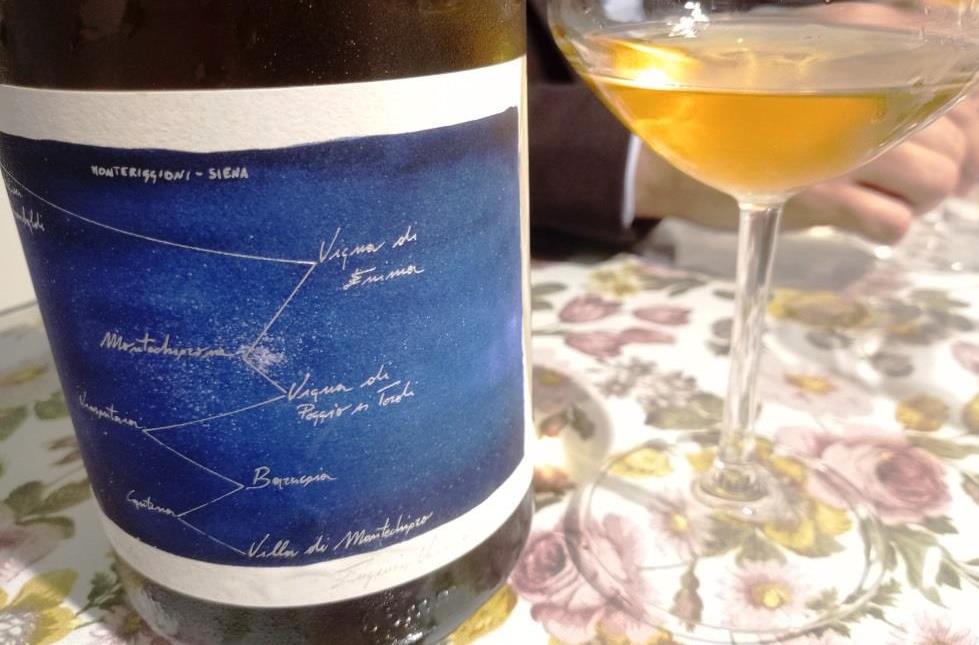 Wine bottle with a blue label featuring a handwritten constellation-style map next to a wine glass on a floral tablecloth. The setting appears to be a casual wine tasting or dining scene.