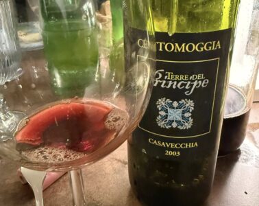 Bottle of wine with a dark label reading Casavecchia 2003, placed beside a glass on a table; a white flower is visible in the background near the bottle.