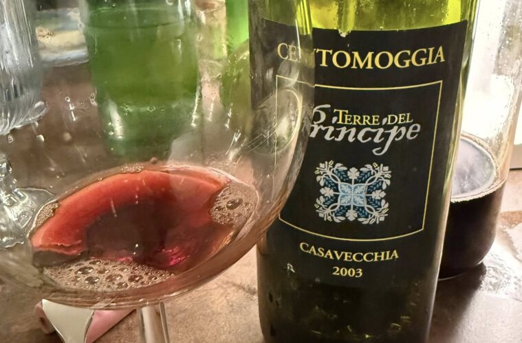 Bottle of wine with a dark label reading Casavecchia 2003, placed beside a glass on a table; a white flower is visible in the background near the bottle.
