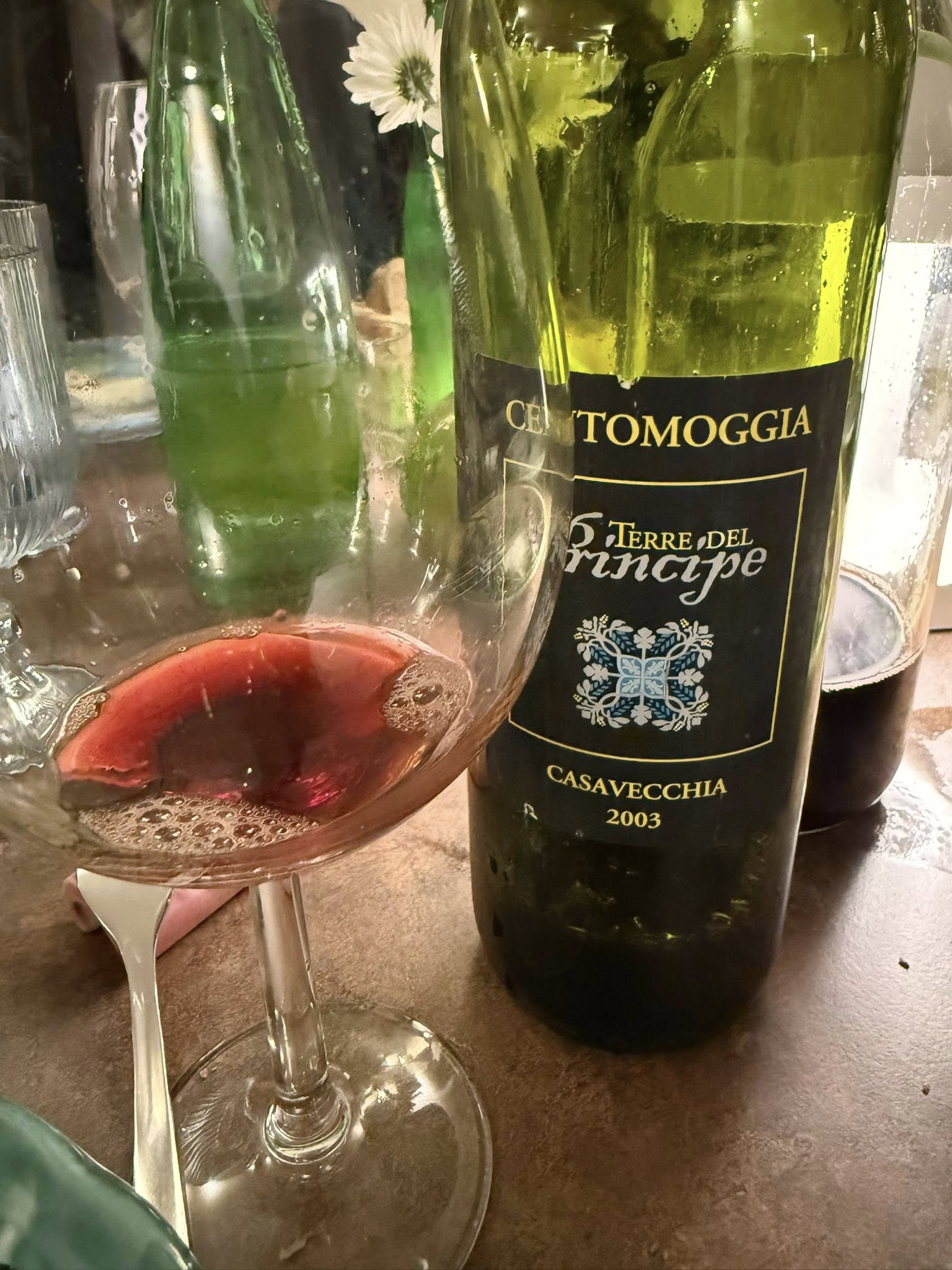 Bottle of wine with a dark label reading Casavecchia 2003, placed beside a glass on a table; a white flower is visible in the background near the bottle.