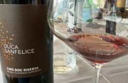 Bottle of Duca Sanfelice Cirò Doc Riserva red wine beside a filled wine glass on a table set with a water bottle in the background.