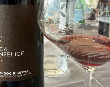 Bottle of Duca Sanfelice Cirò Doc Riserva red wine beside a filled wine glass on a table set with a water bottle in the background.