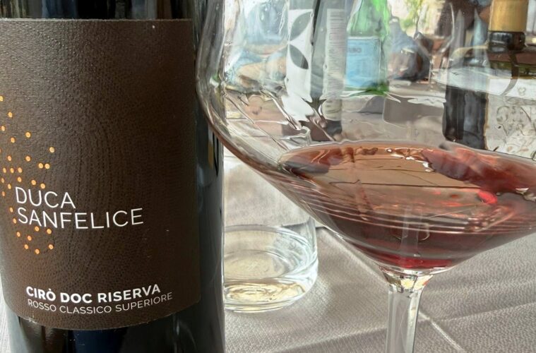 Bottle of Duca Sanfelice Cirò Doc Riserva red wine beside a filled wine glass on a table set with a water bottle in the background.
