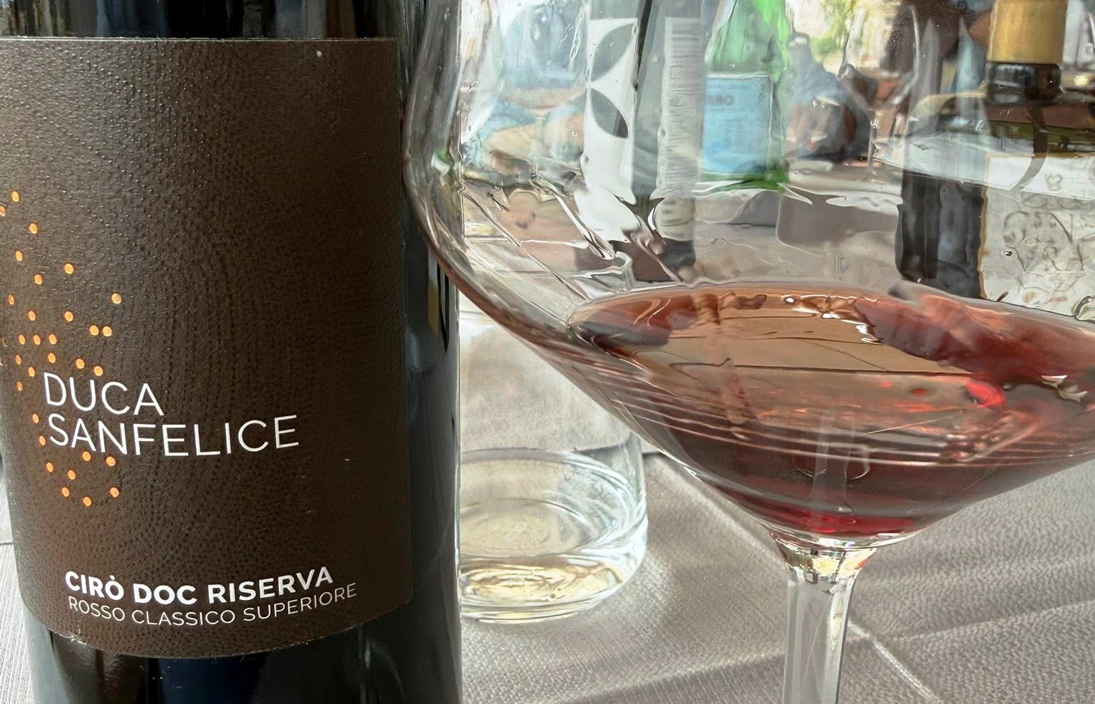 Bottle of Duca Sanfelice Cirò Doc Riserva red wine beside a filled wine glass on a table set with a water bottle in the background.