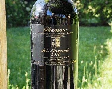 Dark glass wine bottle outdoors with a black label showing 'Ghemme dei Mazzeni 2010' in a grassy garden setting.