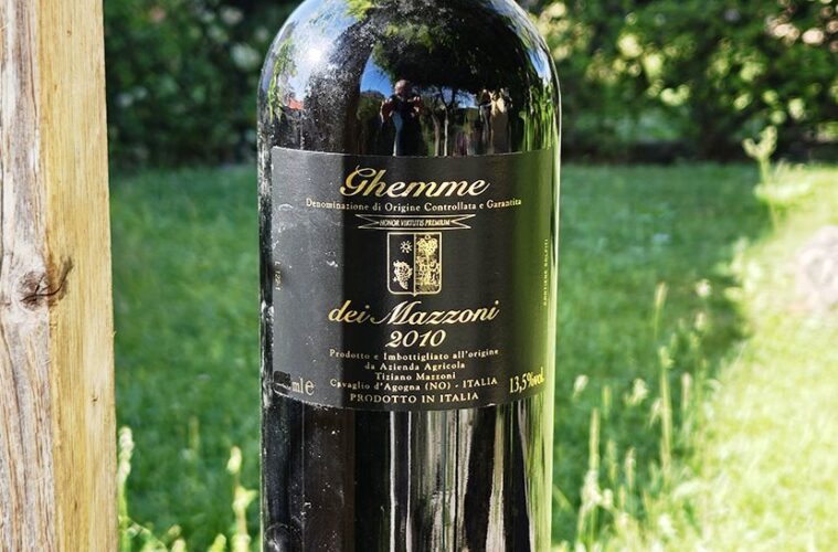 Dark glass wine bottle outdoors with a black label showing 'Ghemme dei Mazzeni 2010' in a grassy garden setting.