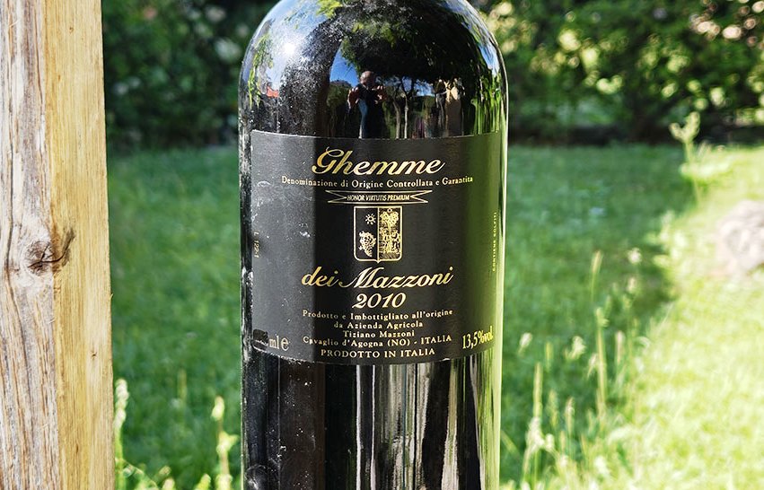 Dark glass wine bottle outdoors with a black label showing 'Ghemme dei Mazzeni 2010' in a grassy garden setting.