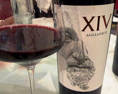 Red wine glass in the foreground with a bottle labeled XIV Aglianico and a sketchy artwork on the label in the background.