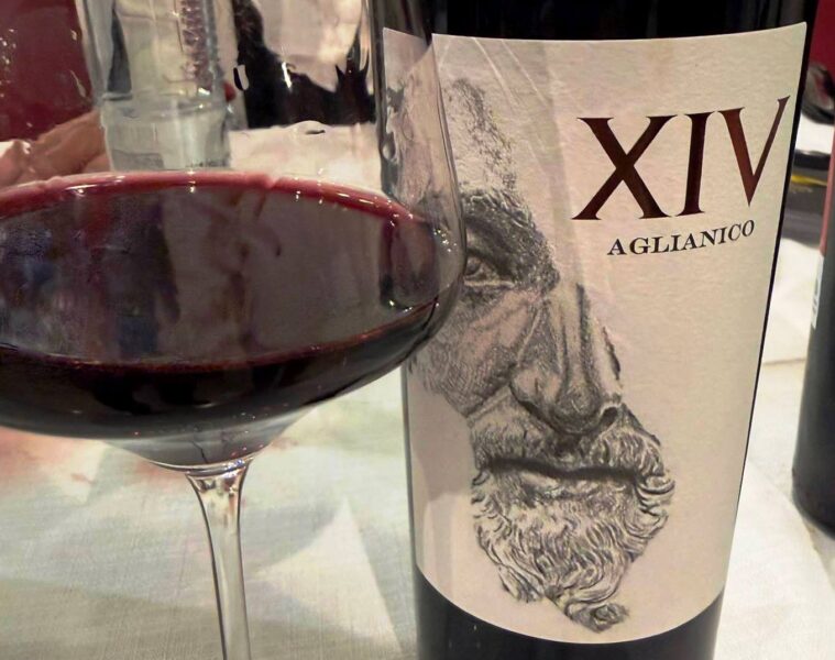 Red wine glass in the foreground with a bottle labeled XIV Aglianico and a sketchy artwork on the label in the background.