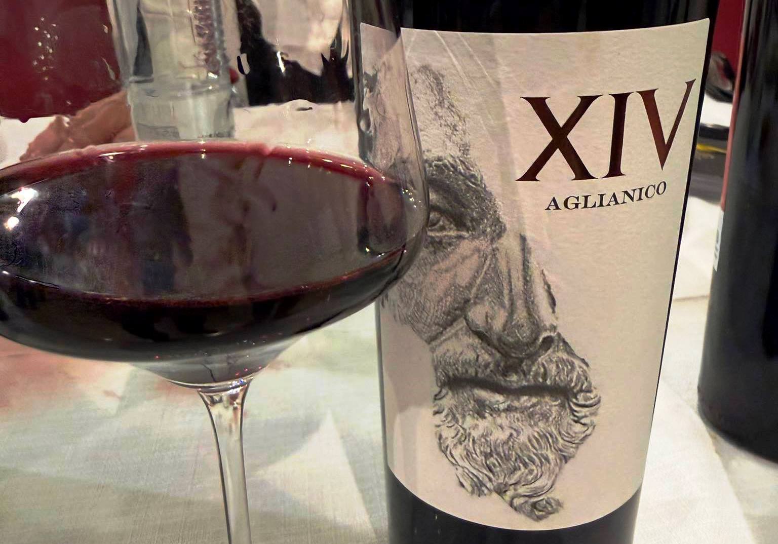 Red wine glass in the foreground with a bottle labeled XIV Aglianico and a sketchy artwork on the label in the background.