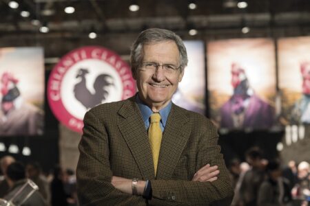 Smiling older man in a brown blazer and yellow tie stands with arms crossed at a conference, with banners and attendees in the blurred background.