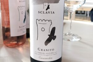 Bottle of Sclavia Granito wine with white label and black brushstroke design, on a white table beside a wine glass.