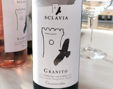 Bottle of Sclavia Granito wine with white label and black brushstroke design, on a white table beside a wine glass.