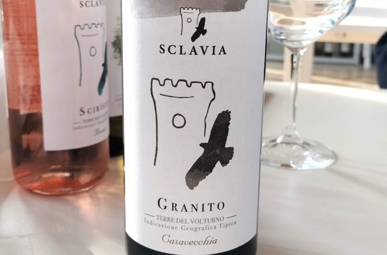 Bottle of Sclavia Granito wine with white label and black brushstroke design, on a white table beside a wine glass.