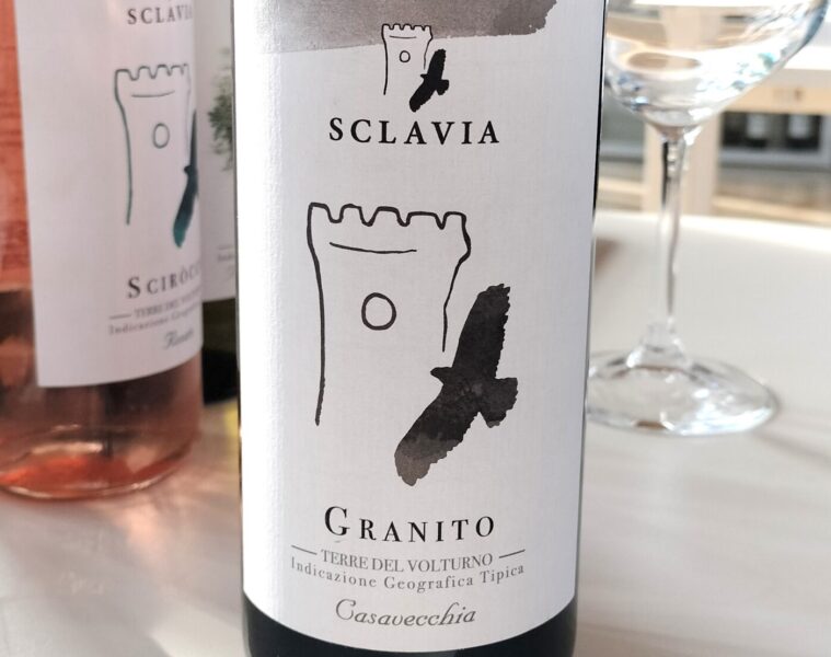 Bottle of Sclavia Granito wine with white label and black brushstroke design, on a white table beside a wine glass.