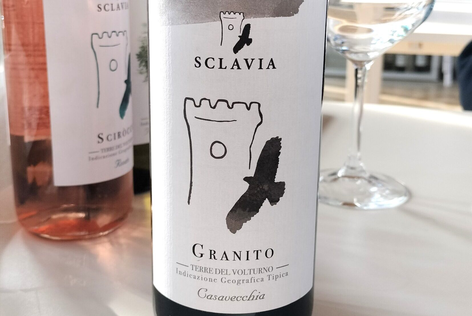 Bottle of Sclavia Granito wine with white label and black brushstroke design, on a white table beside a wine glass.