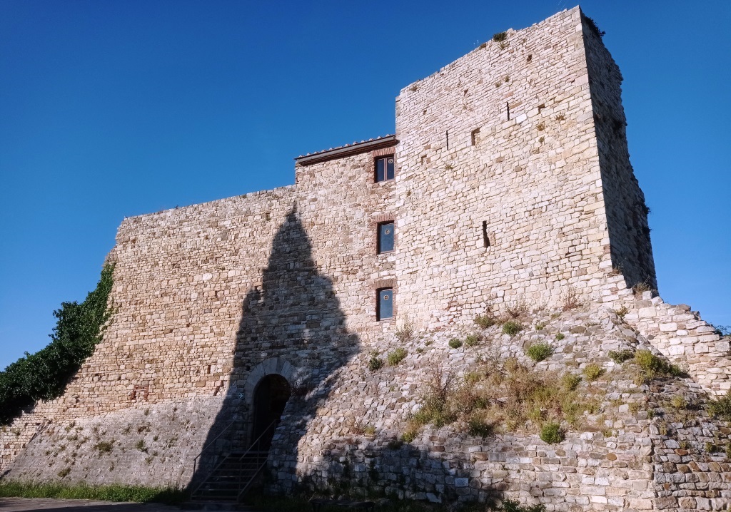 Stone fortress on a hill under a clear blue sky; a tall wall casts a dark shadow across the lower stones.