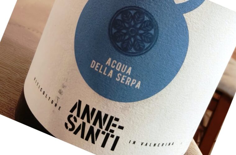 Close-up of a bottle label with a blue circular emblem and the text 'ACQUA DELLA SERPA' on a light background. The label sits on a wooden surface.