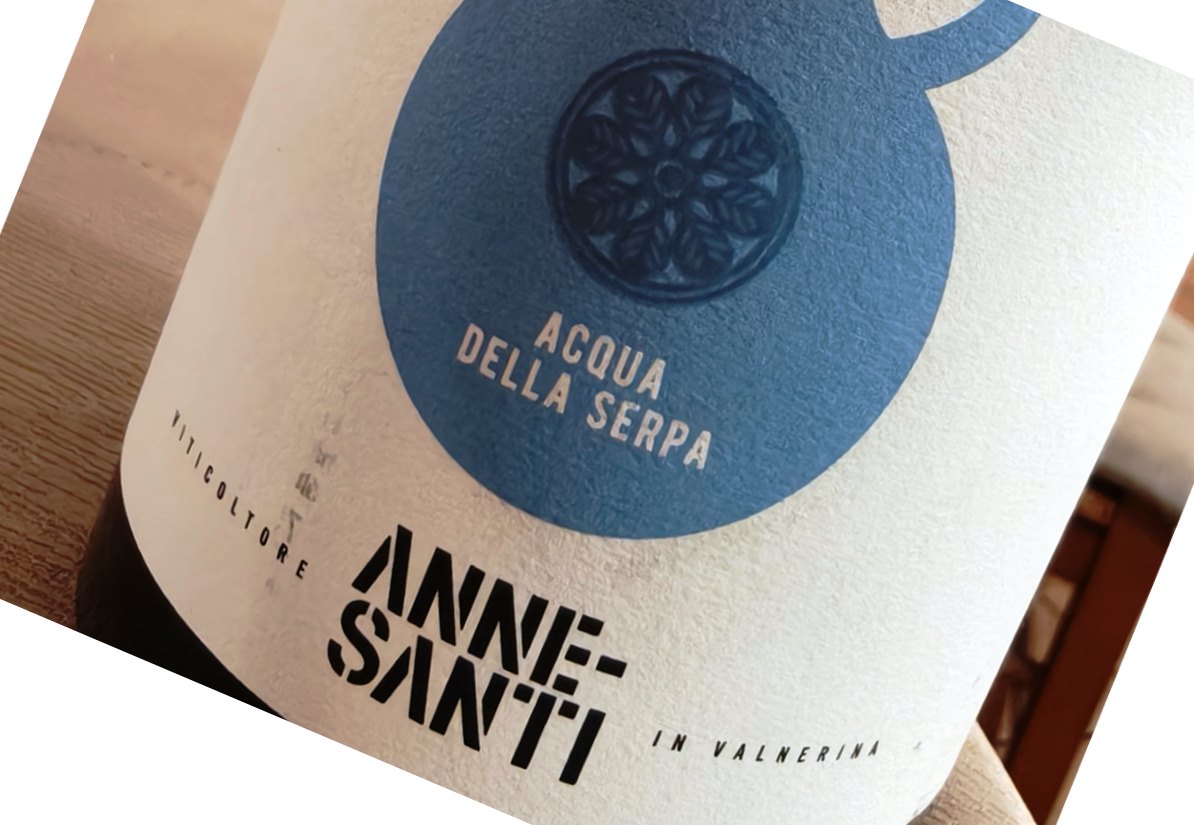 Close-up of a bottle label with a blue circular emblem and the text 'ACQUA DELLA SERPA' on a light background. The label sits on a wooden surface.