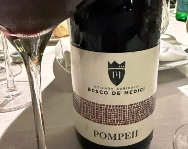 Wine bottle with label reading Bosco De Medici Pompeii on a dining table with wine glasses nearby.