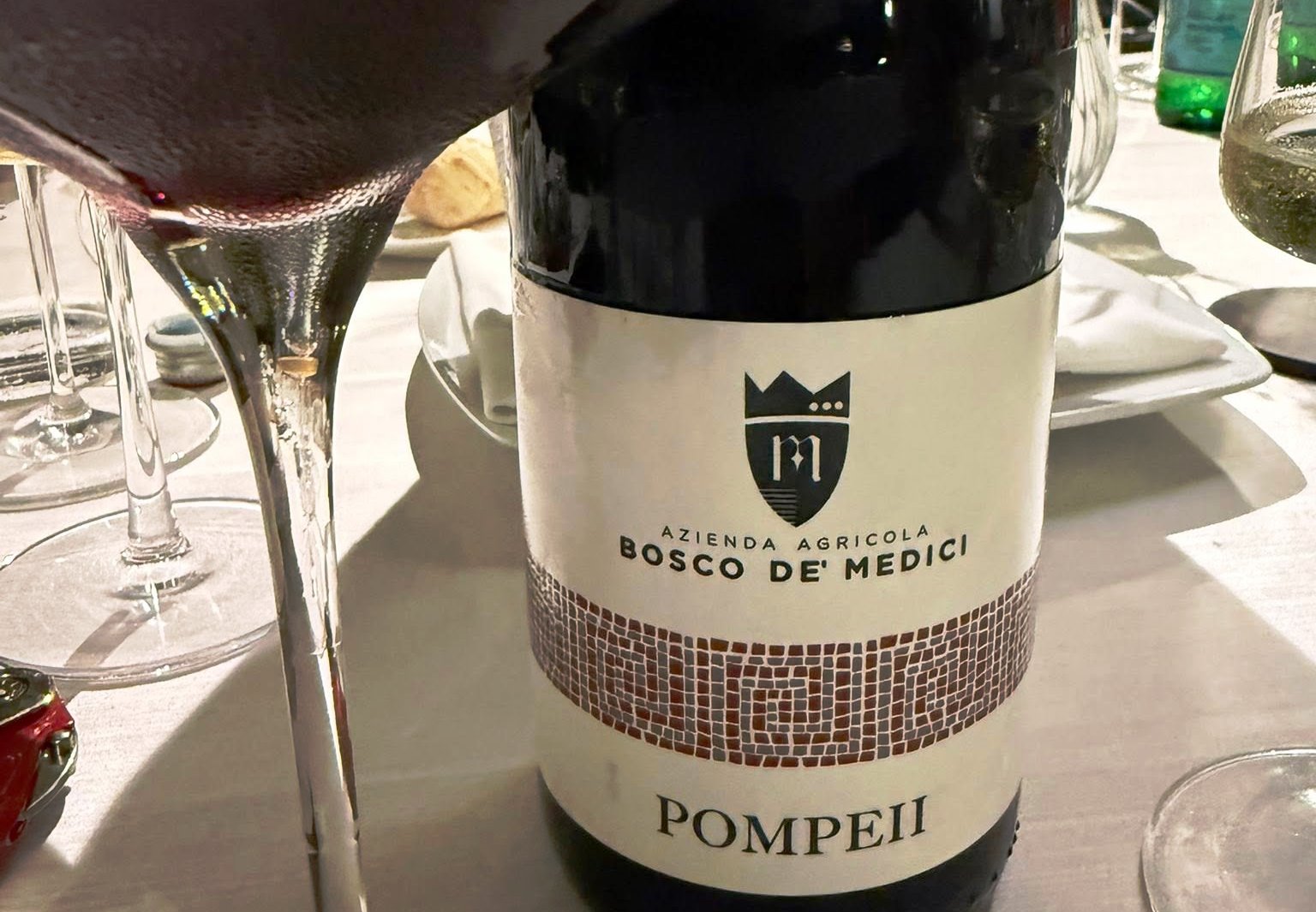 Wine bottle with label reading Bosco De Medici Pompeii on a dining table with wine glasses nearby.