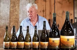 Older man in a light blue shirt sits behind a row of wine bottles on a wooden table, likely at a tasting or presentation, with a rustic backdrop.