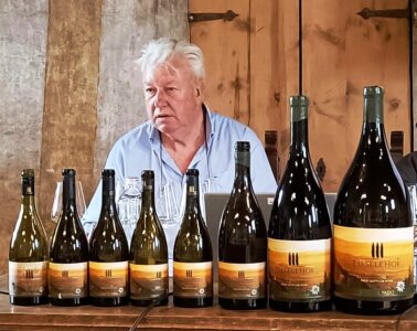 Older man in a light blue shirt sits behind a row of wine bottles on a wooden table, likely at a tasting or presentation, with a rustic backdrop.