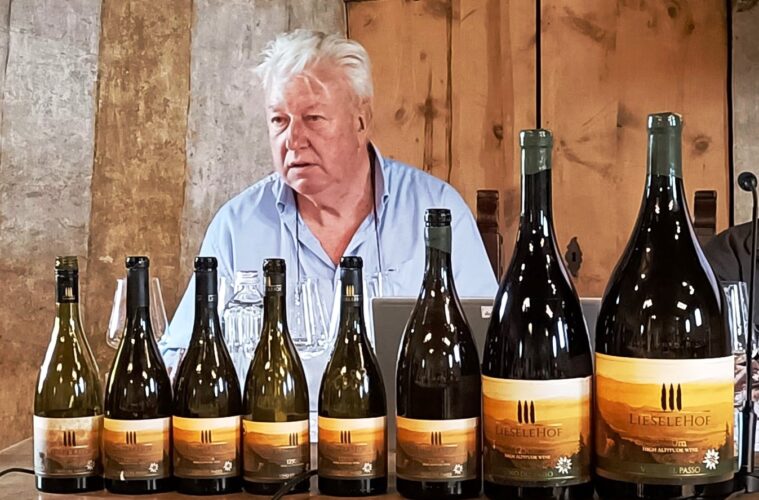 Older man in a light blue shirt sits behind a row of wine bottles on a wooden table, likely at a tasting or presentation, with a rustic backdrop.