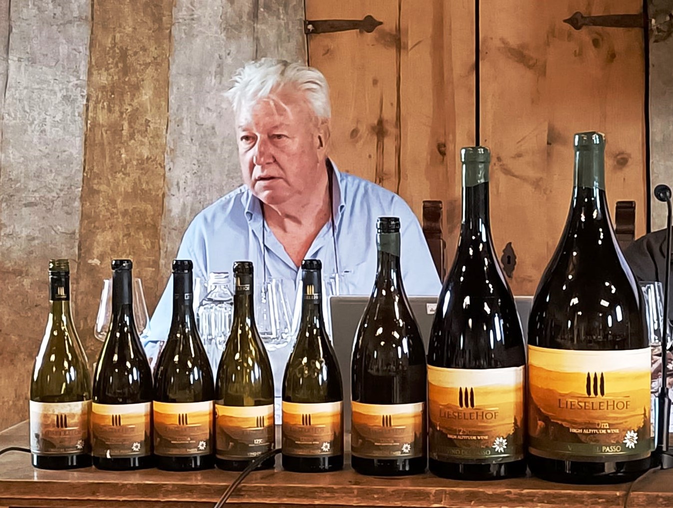 Older man in a light blue shirt sits behind a row of wine bottles on a wooden table, likely at a tasting or presentation, with a rustic backdrop.