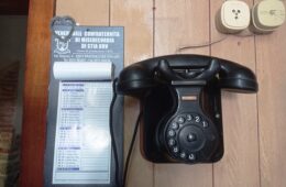 Vintage black rotary telephone on a wooden desk beside a blue plaque with Italian text and a heart-shaped metal ornament.