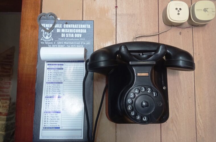 Vintage black rotary telephone on a wooden desk beside a blue plaque with Italian text and a heart-shaped metal ornament.