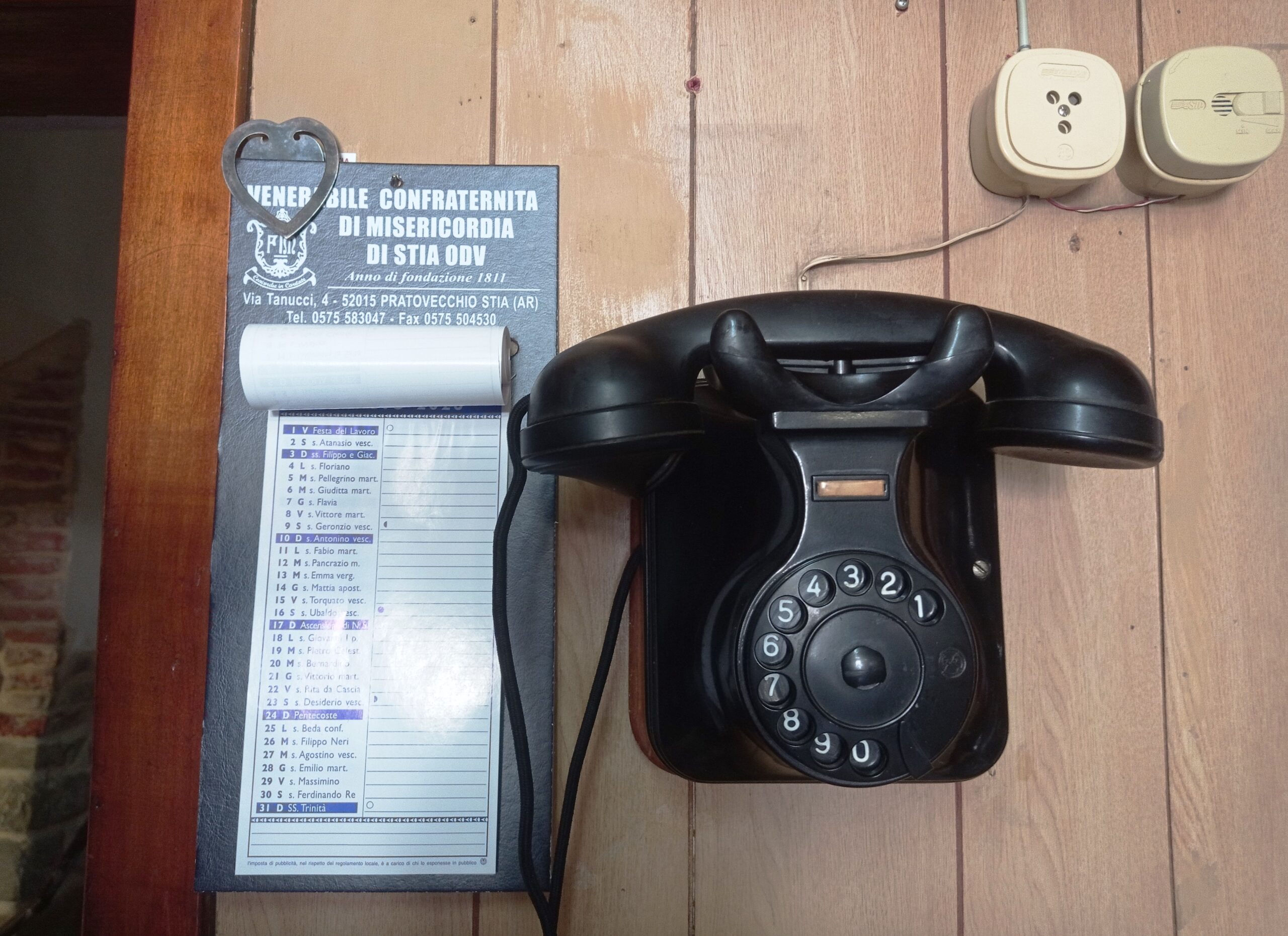 Vintage black rotary telephone on a wooden desk beside a blue plaque with Italian text and a heart-shaped metal ornament.