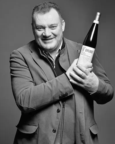 Smiling man in a blazer holds a wine bottle toward the camera in a grayscale photo.
