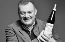 Smiling man in a blazer holds a wine bottle toward the camera in a grayscale photo.