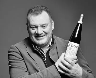 Smiling man in a blazer holds a wine bottle toward the camera in a grayscale photo.