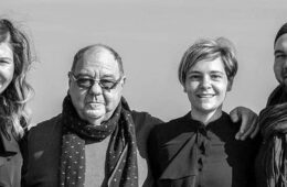Four adults posing together outdoors, smiling at the camera, with arms around each other in a line.